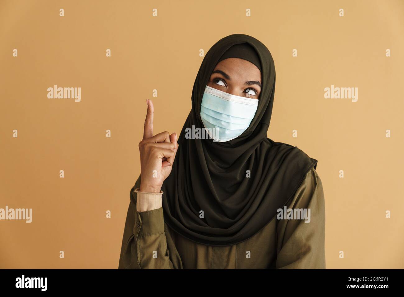 black-muslim-woman-in-face-mask-looking-and-pointing-finger-upward-isolated-over-beige-backgro...jpg